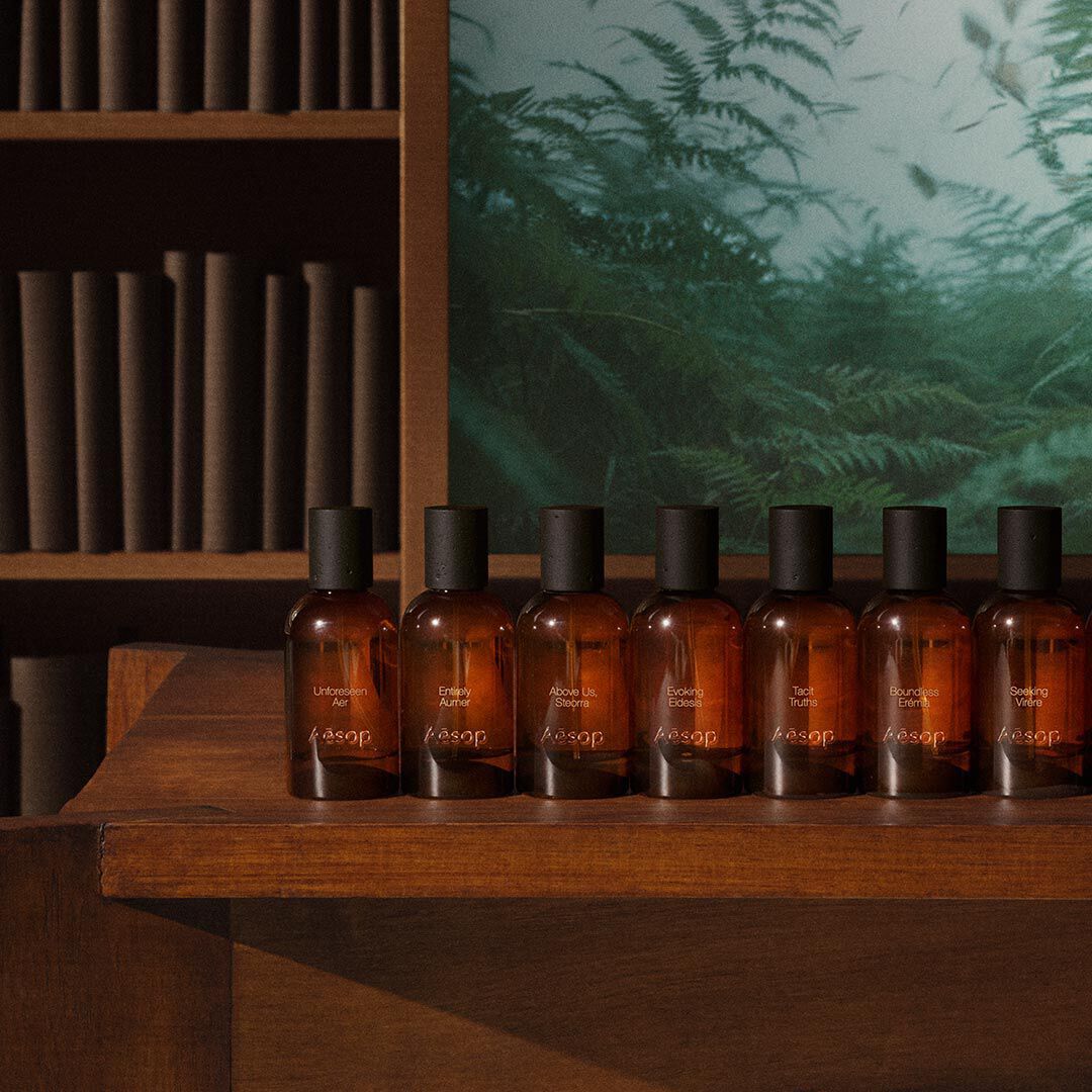 Row of amber Aesop fragrance bottles on a wooden table beside shelves and a green fern image.